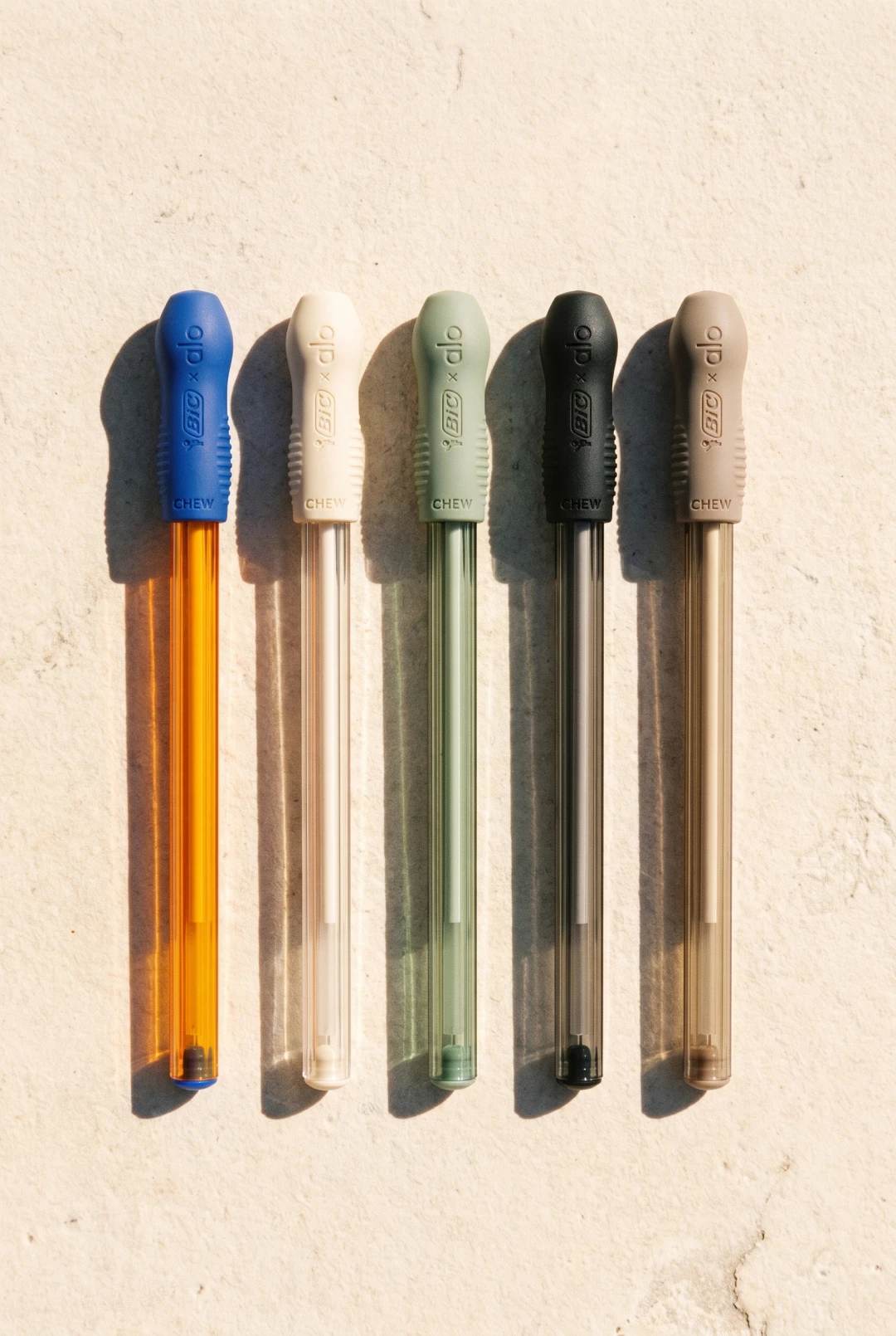 Cinco lapiceras alineadas con capuchones azul, crudo, verde salvia, negro y beige grabados BiC x alo CHEW, sobre una pared de cemento claro con sombras